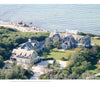 An aerial view from "United by Design" by Schiffer Publishing, Ltd. shows two large gray-shingled Cape Cod homes, set among greenery and landscaped lawns, with a driveway and shoreline waves in the background.