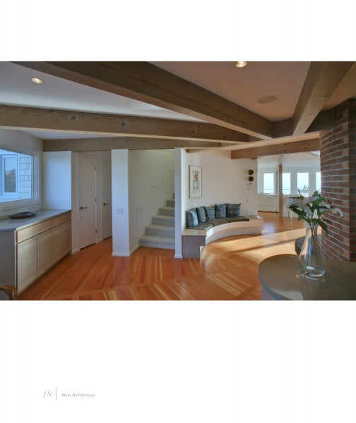 United by Design by Schiffer Publishing, Ltd. captures the airy charm of Nantucket homes with wooden beams, light wood floors, a curved staircase, built-in blue seating, large windows, and a brick column for a bright, open space.