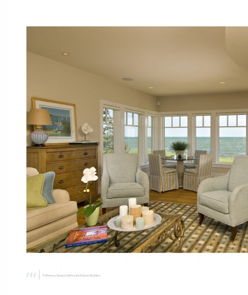 A cozy living room inspired by Martha’s Vineyard features armchairs, a sofa, and a striped rug. The table displays “United by Design” by Schiffer Publishing, Ltd., while large windows offer an ocean view and a nearby striped dining area.