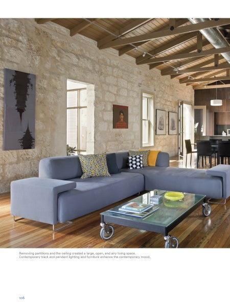 A modern King William living room, as seen in "Old Places, New Spaces" by Schiffer Publishing, Ltd., features a gray sectional, vibrant pillows, glass coffee table on wheels, art on stone walls, wood floors, and exposed beams leading to the dining area.