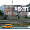 A yellow taxi passes a weathered brick building featuring Detroit Graffiti by Schiffer Publishing, Ltd.—large white NEKST and smaller gray PHANG—while overgrown plants line the concrete barrier, enhancing the street art scene.