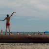 A person in red shorts stands on a large pipe at the beach, embodying New York surfing as seen in "Surf NYC" by Schiffer Publishing, Ltd., with people, umbrellas, and the Atlantic Ocean in the partly cloudy background.