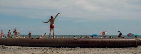 A person in red shorts stands on a large pipe at the beach, embodying New York surfing as seen in "Surf NYC" by Schiffer Publishing, Ltd., with people, umbrellas, and the Atlantic Ocean in the partly cloudy background.