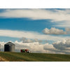 Two metal grain silos and a red farm truck on a grassy hill under dramatic clouds evoke scenes from "Yellowstone Country" by Schiffer Publishing, Ltd., with a dirt road winding through the picturesque landscape.