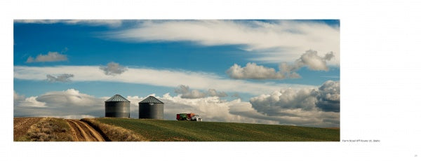 Two metal grain silos and a red farm truck on a grassy hill under dramatic clouds evoke scenes from "Yellowstone Country" by Schiffer Publishing, Ltd., with a dirt road winding through the picturesque landscape.