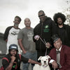 Six men pose outside with a white dog; five wear casual attire while one, evoking 1980s musicians, sits in front in a suit and hat. Trees and buildings stand beneath an overcast sky. Featured: "80s Redux" by Schiffer Publishing, Ltd.