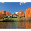 A row of houses on a grassy hill amid orange and yellow autumn trees reflects in a calm lake—an idyllic landscape featured in "New Hampshire" by Schiffer Publishing, Ltd., perfect for scenic photography lovers.