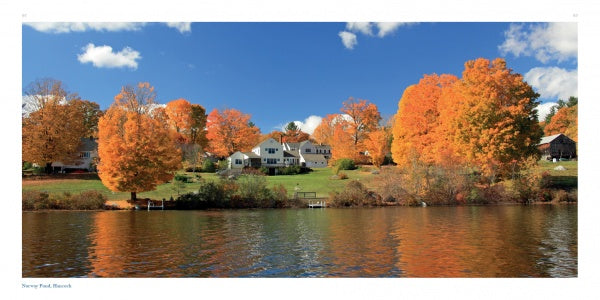 A row of houses on a grassy hill amid orange and yellow autumn trees reflects in a calm lake—an idyllic landscape featured in "New Hampshire" by Schiffer Publishing, Ltd., perfect for scenic photography lovers.