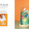 The Little Paper Universes kit by Schiffer Publishing, Ltd. features an open book with jungle-themed 3-D paper art displayed under a glass dome on a wooden base. One page shows crafting steps; the other showcases the finished project.