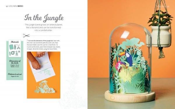 The Little Paper Universes kit by Schiffer Publishing, Ltd. features an open book with jungle-themed 3-D paper art displayed under a glass dome on a wooden base. One page shows crafting steps; the other showcases the finished project.