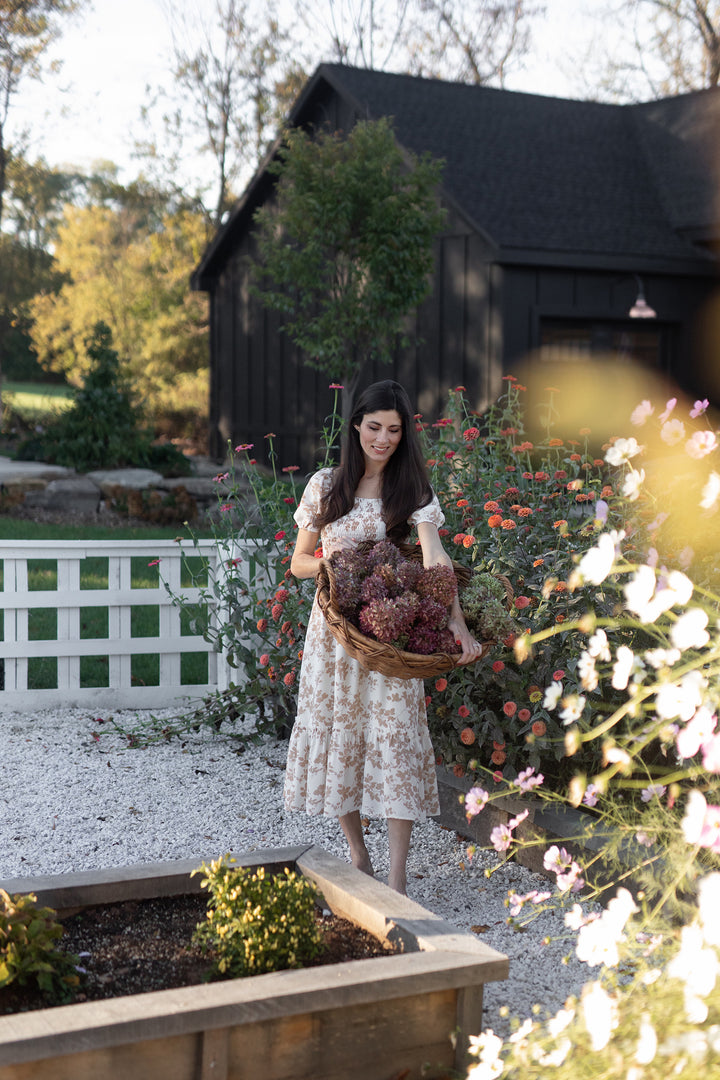 A woman in a floral dress stands in a garden holding dried hydrangeas—a scene from "Modern HomeMaker" by Schiffer Publishing, Ltd., perfect for creative homeowners seeking home décor inspiration.