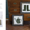 Two square textile artworks from Schiffer Publishing, Ltd.'s Rug Hooking with Wool Strips feature green boots and a watering can. Displayed on terra-cotta tiles with gardening tools, they create a rustic, cozy look.