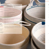 Several coiled cotton rope baskets in neutral colors and different sizes, some with handles, are grouped together. To the right is a table of contents for "Artisan Ropework" by Schiffer Publishing, Ltd., listing its chapters and pages.