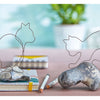 Two Whimsical Wire squirrel sculptures by Schiffer Publishing, Ltd. sit on rocks atop notebooks with colored pencils and shavings nearby. A green plant is blurred in the background—perfect for those new to wire crafts.