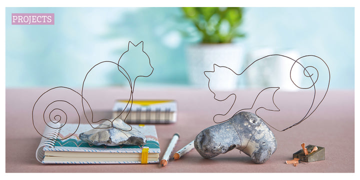 Two Whimsical Wire squirrel sculptures by Schiffer Publishing, Ltd. sit on rocks atop notebooks with colored pencils and shavings nearby. A green plant is blurred in the background—perfect for those new to wire crafts.