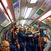 A photo from METRO / New York / London / Paris by Schiffer Publishing, Ltd. shows a crowded London Tube train with passengers, some standing and holding red poles, others sitting—many appear tired. A loneliness quote appears on the image's right side.