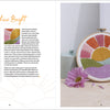 An open Sunny Stitches book by Schiffer Publishing, Ltd. shows a project guide on the left and a beginner sunset, mountains, and fields embroidery hoop art on the right, propped by a pink daisy on a light wood surface.