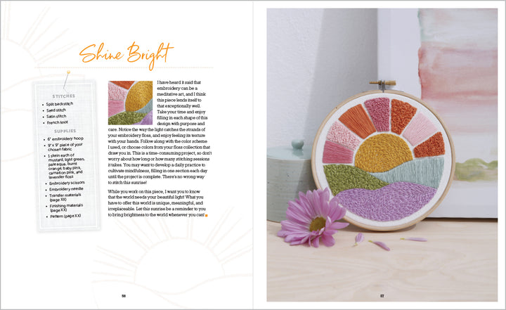 An open Sunny Stitches book by Schiffer Publishing, Ltd. shows a project guide on the left and a beginner sunset, mountains, and fields embroidery hoop art on the right, propped by a pink daisy on a light wood surface.