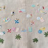 Close-up of the Juno's Nature Embroidery Notebook by Schiffer Publishing, Ltd., featuring playful beach-themed and nature embroidery patterns with people, palm trees, umbrellas, and accessories on beige fabric.