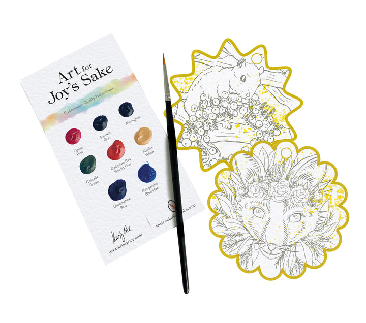 The Paint-Your-Own Holiday Decorations kit by Schiffer Publishing, Ltd. features a palette card, thin brush, and two animal-themed ornaments—a rabbit with flowers and a fox with a floral crown—on a white background.