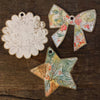 Three wooden ornaments from Schiffer Publishing’s Paint-Your-Own Holiday Decorations: a scalloped circle with an animal face, a floral-patterned bow, and a star with botanical detail, inspired by Kristy Rice’s watercolor holiday designs.