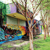 The mural "Arte del Pueblo" by Schiffer Publishing, Ltd. displays vibrant abstract animals on a building’s side in San Antonio, with sunlight streaming through nearby trees and casting patterned shadows on the sidewalk and grass below.