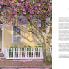 A blooming pink cherry blossom tree adorns a yellow house with white trim and a picket fence, creating an idyllic scene from Nantucket by Schiffer Publishing, Ltd., with petals scattered across the brick path and porch.