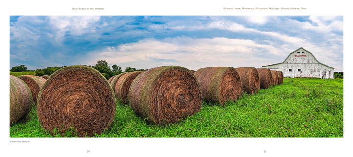 Large round hay bales rest in a green field beneath a cloudy sky, with a white barn behind. "Back Roads of the Midwest" by Schiffer Publishing, Ltd. showcases David Skernick’s peaceful rural landscape photography.