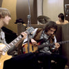 Two musicians play electric guitars and a drummer is in the background behind brown sound barriers in a casual recording studio, reminiscent of Fleetwood Mac’s blues era—just like "Fleetwood Mac in Chicago" from Schiffer Publishing, Ltd.