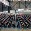 Hundreds in dark suits line up inside a hangar with a Japan Airlines plane and a screen behind—a striking image from Japan by Schiffer Publishing, Ltd., perfect for any travel photography collection.