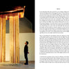 A man stands beneath "Hammer and Tongs" by Schiffer Publishing, Ltd., its vertical beams and rusted metal plate displayed on round stones in an art gallery—a striking example of public sculpture.