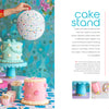A vibrant party scene showcases three cakes on pastel stands, confetti, a hand with a white paper lantern, and blue floral wallpaper, with text about a DIY cake stand project from Schiffer Publishing, Ltd.'s The Big Book of Happy Crafts.